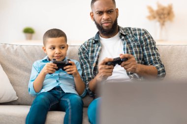 Genç Siyah Baba ve Küçük Oğul video oyunları oynuyorlar. Evde oyun alanı kontrolörleriyle oturuyorlar. Aile Bağları Eğleniyor, Baba ve Çocuk Hafta Sonu Birlikte Video Oyunları Oynuyor