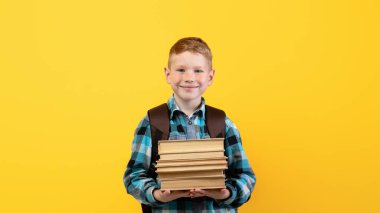 Sırt çantalı gülümseyen okul çocuğu bir sürü kitap tutuyor, sarı stüdyo arka planı, fotokopi odası. Mutlu çocuk okula gidiyor, kitap tutuyor, okul günlerinden zevk alıyor. Çocuk eğitimi konsepti