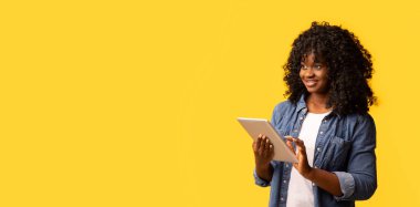 En yeni dijital tabletle neşeli Afro-Amerikalı kadın sarı stüdyo arkaplanındaki fotokopi alanına bakıyor ve gülümsüyor, panorama. Mobil uygulama konsepti reklamı