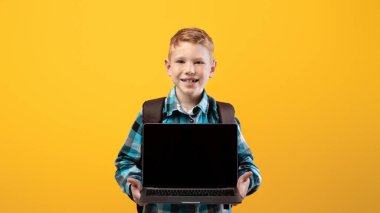 Cheerful ginger kid with schoolbag showing laptop with blank screen on yellow studio background, copy space. Happy schoolboy holding modern laptop, using technologies in education, mockup