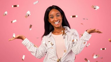 Tatil satışı için seçenekler. Neşeli siyahi kadın varyantları, reklam ürünlerini karşılaştırıyor, pembe stüdyo arka planında görünmez nesneleri tutuyor ve düşen konfetilerle, tasarım için modelleme yapıyor.