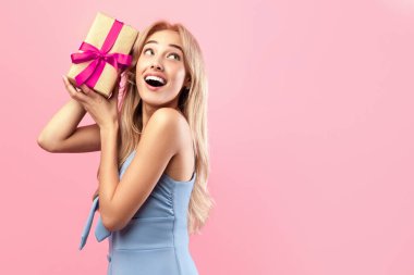 Şirin bir elbise içinde, hediye paketini sallayan beyaz bir kadın pembe stüdyo arka planında çıngırağı dinliyor, fotokopi alanı olan afiş tasarımı yapıyor. Tatil kutlaması konsepti
