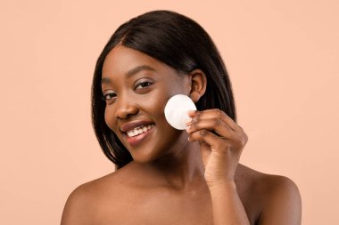 Çekici genç siyahi kadın havluya sarılıp tonerle yüzünü temizliyor, pamuk ped ve yüz ürünü kullanıyor, yakın plan, bej renkli stüdyo arka plan. Yüz bakımı, yüz temizleme konsepti