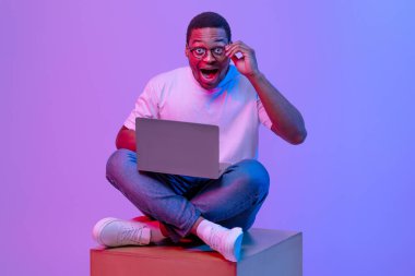 Küpte dizüstü bilgisayarlı, mor ışıklı, ağzı açık bir şekilde kameraya bakan Afro-Amerikan erkek öğrencinin portresi eğitim için modern teknoljilerin keyfini çıkarıyor.