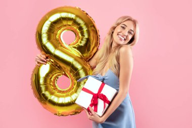 Kadınlar Günü kutlama konsepti. Altın rengi balonlu ve hediye kutulu çekici bayan kameraya bakıyor ve pembe stüdyo arka planında gülümsüyor.