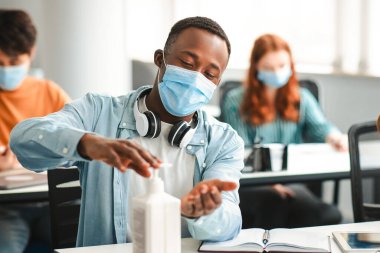 Temizlik ve Koruma Konsepti. Siyahi öğrenci şişeden elde antibakteriyel dezenfektan uyguluyor, ameliyat maskesi takıyor, sınıfta oturuyor. Enfeksiyon ve mikropların yayılmasını önler.