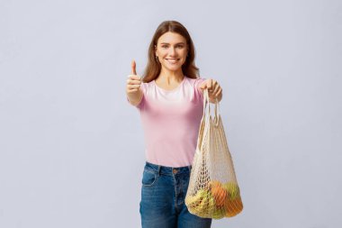 Bir kadın kendine güvenerek duruyor, elinde meyve dolu bir ağ torbası tutarken başparmağını kaldırıyor. Günlük kıyafetleri ve olumlu tavırları, çevre dostu uygulamalara olan bağlılığını vurguluyor..