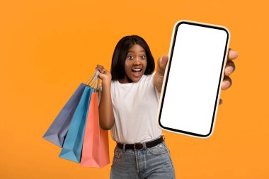 Satış sezonu. Alışveriş torbaları ile turuncu stüdyo geçmişini gösteren hayret verici genç siyahi bayan. Afro-Amerikan kadının elinde alışverişler var, fotokopi alanına bakıyor ve el kol hareketi yapıyor.