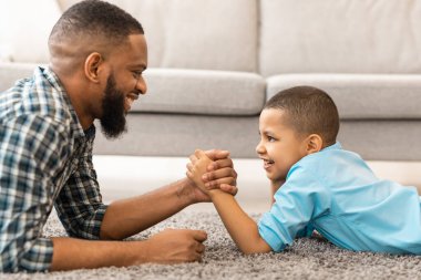 Mutlu Afrika kökenli Amerikalı baba ve oğul bilek güreşi yapıyorlar. Evde yerde yatıyorlar. Mutlu Genç Baba ve Küçük Oğlan Kol Güreşi Birlikte Eğleniyor. Yan Görünüm