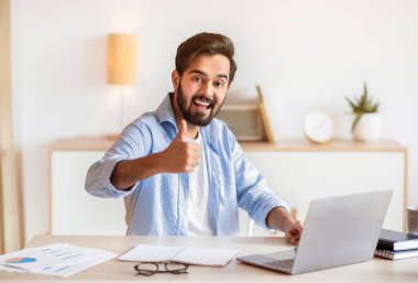 Uzak İş Fırsatları. Laptopta çalışan ve kamerada el hareketlerini gösteren mutlu Arap serbest yazar, doğulu adam uzaktan çalışmaktan hoşlanıyor, bilgisayarla masada oturuyor, boş yer var.