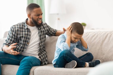 Kızgın Afrikalı baba evdeki koltukta oturan korkmuş oğlana bağırıyor. Öfkeli baba ona bağırırken mutsuz çocuk ağlayan kulakları kapatıyor. Çocuklara karşı aile içi şiddet. Seçici Odaklanma