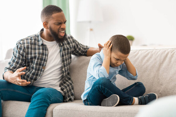 Злой африканский отец кричит на испуганного сына, сидящего дома на диване. Несчастный мальчик плачет, закрывая уши, в то время как разъяренный отец кричит на него. Домашнее насилие в отношении детей. Селективный фокус