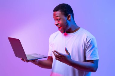 Çevrimiçi teklif. Şaşırmış Afrikalı Amerikalı adam elinde dizüstü bilgisayarla parlayan ekrana bakıyor, heyecanlı siyahi adam oyunlar oynuyor ya da bilgisayarda internete takılıyor, Mor Arkaplanda Neon Işığı altında mahsur kalıyor.