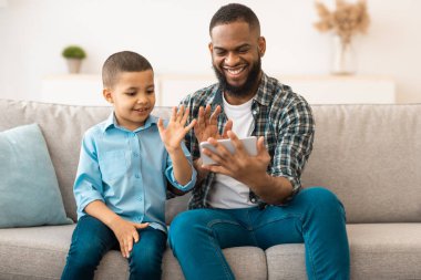 Siyah Baba ve Oğlu, akıllı telefondan cep telefonuna el sallayarak evdeki koltukta otururken görüntülü arama yapıyor. Telefon Kavramı için Uzaktan İletişim Uygulaması