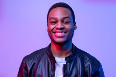Neon ışığı altında deri ceketle poz veren mutlu siyah adam portresi, yakışıklı Afro-Amerikan erkeği mor menekşe arka planının başında dikilirken ve gülümserken,