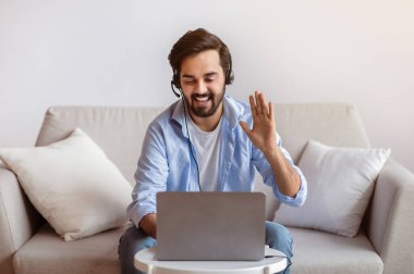 Dost canlısı Doğulu adam evde dizüstü bilgisayarla video görüşmesi yapıyor, web kamerasına el sallıyor, birini selamlıyor. Oturma odasında koltukta otururken internetten haberleşmenin keyfini çıkaran genç bir Arap. Boşluğu kopyala.