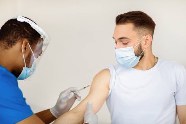 Genç beyaz adam hastanede koronavirüs aşısı yaptırıyor. Afrikalı Amerikalı bir tıp çalışanı erkek hastayı klinikte kovid 19 'a karşı bağışık hale getiriyor. Sağlık ve küresel enfeksiyon önleme
