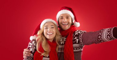 Mutlu genç adam ve kız arkadaşı Noel kıyafetleri içinde kırmızı arka planda selfie çekiyorlar.