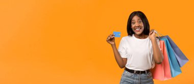 Sınırsız alışveriş konsepti. Uzun saçlı, neşeli Afro-Amerikalı kadın elinde renkli alışveriş çantaları var. Kredi kartı harcamaları turuncu stüdyo arka planında, sahtesinde fotokopi alanı olan bir panorama.