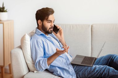 Arap adam evde dizüstü bilgisayar ve cep telefonuyla çalışırken sorun yaşıyor, Doğulu serbest çalışan adam internet bağlantısıyla sorun yaşıyor, müşteri hizmetleri müdürüyle konuşuyor, koltukta oturuyor.