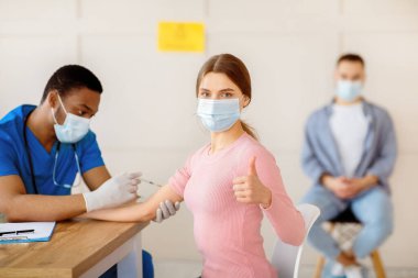 Tıbbi maskeli genç bir kadın kovid 19 aşısını onaylıyor, klinikte baş parmak hareketi gösteriyor. Milenyum bayanı sağlık merkezinde korovirüs aşısı öneriyor.