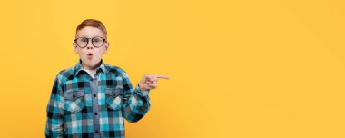 Gözlüklü hayret verici bir çocuk, mesaj ya da reklam için fotokopi alanını hedefliyor. Şaşırmış kızıl saçlı okul çocuğu sarı stüdyo arka planındaki boşluğu işaret ediyor, panorama