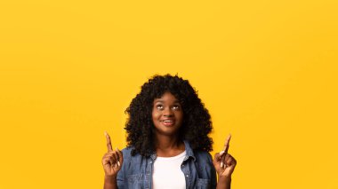 Afrikalı Amerikalı kıvırcık kadın sarı stüdyo arka planında fotokopi alanını işaret ediyor. Genç ve güzel siyahi bayan gülümsüyor ve mesaj ya da reklam için boş alana işaret ediyor.