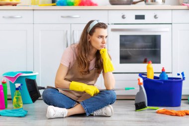 Mutsuz milenyum hizmetçisi mutfak zemininde bir sürü malzemeyle oturuyor. Temizlikten bıkıp usanmış. Önlüklü ve lastik eldivenli genç bir kadın ev işinden yorgun düşmüş.