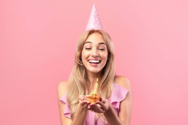 Şirin elbiseli neşeli sarışın kadın portresi ve pembe stüdyo arka planında mumlu doğum günü çöreği. Kalp şeklinde balonu olan, neşeli tatlı yiyen mutlu bir milenyum kadını.