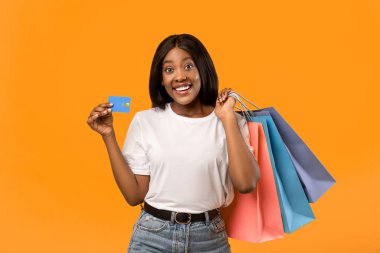 Alışveriş torbaları ve kredi kartıyla turuncu stüdyo arka planında gülümseyen mutlu siyah kadın, boş alan. Afro-amerikan kadın alışveriş kolikleri ve limitsiz kredi kartları.