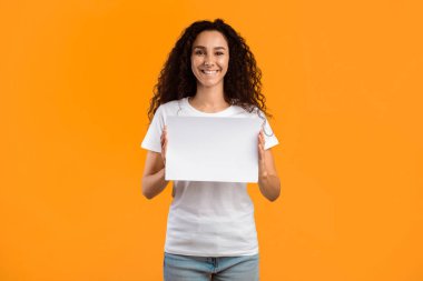 Happy Lady Holding Empty Paper Board Smiling To Camera Standing Over Yellow Studio Background. Lady Showing Blank Poster Advertising Your Text. Mockup