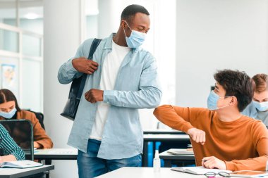 Virüs Kavramını yaymayı bırak. Birbirlerini selamlayan ve sınıfta dirsek tokuşturan koruyucu maskeler takan gülümseyen öğrenciler. Lisede okuyan Asyalı ve Afrikalı Amerikalı erkekler