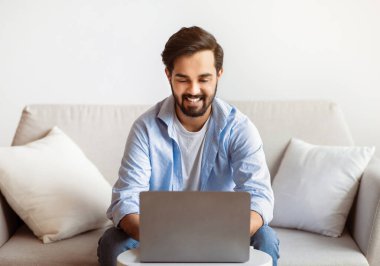 Telekomünikasyon konsepti. Evde dizüstü bilgisayarla uzaktan çalışan neşeli Doğulu Serbest Çalışan Adam, Milenyum Arap 'ı oturma odasında bilgisayar kullanırken, Mesafe İş Fırsatlarının keyfini çıkarırken, Boşluk Kopyalayıcı