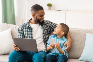 Aile Gadget 'ları. Siyah baba ve küçük oğlu dizüstü bilgisayar ve telefon kullanarak evde oturup sohbet ediyorlar. Baba, Oğlan ve Babası ile Hafta Sonu Bilgisayar ve Akıllı Telefon Birlikte