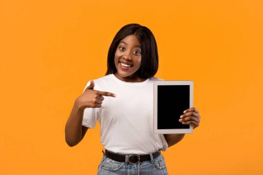 Güzel siyah kadın, boş ekran, turuncu stüdyo arka planı, fotokopi alanı olan yepyeni dijital tableti işaret ediyor. Mutlu Afro-Amerikalı kadın, boş ekranlı, maketli.