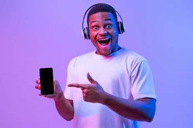 Online Müzik Platformu. Telsiz kulaklıklı heyecanlı Afrikalı adam siyah ekranlı akıllı telefonu işaret ediyor, Mor Arkaplan Üzerinde Neon Işığı Altında Duran Neşeli Milenyum Adamı,