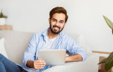 Evinde dijital tabletle gülümseyen genç bir Arap 'ın portresi, Millennial Eastern Guy elinde sekmeli bilgisayarla koltukta oturuyor, kameraya bakıyor, eğlence için modern teknolojilerin tadını çıkarıyor.
