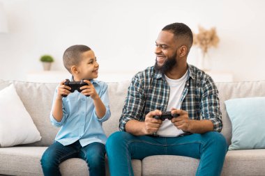 Neşeli Afrikalı Amerikalı baba ve oğul video oyunları oynuyorlar oyun kontrolörleri evde eğleniyor. Hafta sonu tatili konsepti. Genç baba ve çocuğu birlikte bilgisayar oyunu oynuyorlar.