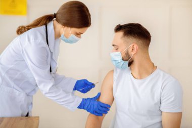 Covid-19 'a karşı aşı. Yüz maskeli kadın doktor, sağlık merkezindeki erkek hastalara koronavirüs aşısı yapıyor. Genç adam klinikte küresel virüse karşı bağışıklık kazanıyor