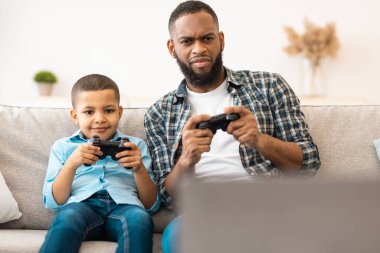 Siyah Baba ve Oğlu video oyunu oynuyorlar. Evdeki koltukta oturup eğleniyorlar. Aile Yaşam Tarzı, Hafta sonu Tatili, Video ve Bilgisayar Oyunları Konsepti. Seçici Odaklanma