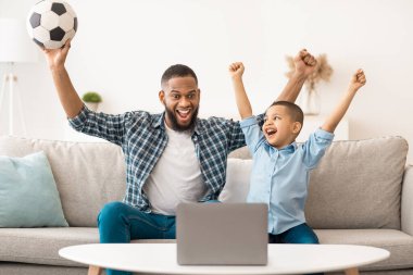 Neşeli Siyah Baba ve Oğlu dizüstü bilgisayarda spor maçı izleyip favori futbol takımını evdeki koltukta oturtuyorlar. Futbol taraftarları, Online Televizyon ve Spor Kanallarını Bilgisayarda İzliyor