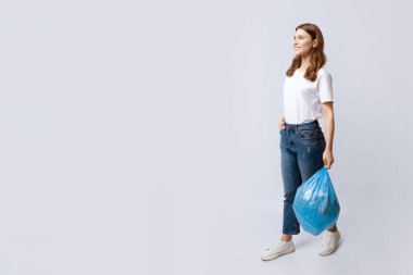 Gülümseyen genç bir ev hanımı, atölyedeki gri arka planda plastik bir çöp torbasıyla yürüyor, olumlu milenyum hanımı, ev atıkları atacak, tam boy çekim yapacak, fotokopi çekecek.