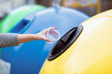 Bir bayan, plastik bardağı canlı ve çevre dostu bir ortamda geri dönüşüm kutusuna koyar. Arka planda sürdürülebilir uygulamaları destekleyen renkli çöp kutuları yer alıyor.