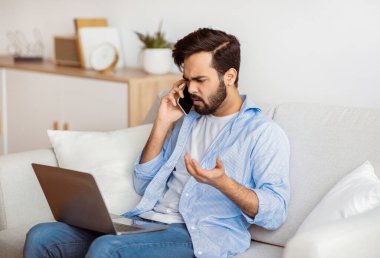 Bilgisayar sorunları. Hüsrana uğramış Arap serbest çalışan adam cep telefonuyla konuşuyor ve evdeki laptop ekranına bakıyor, müşteri yardım hattını arıyor, çevrimiçi çalışırken kötü internet bağlantısı yaşıyor.