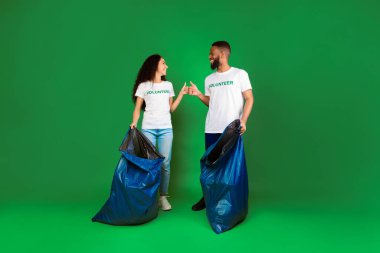 Çevreyi temiz tut. İki kadın ve erkek gönüllü çöp torbaları ile yeşil stüdyonun arka planında çöp topluyor, başparmaklarını kaldırarak el hareketi yapıyorlar. Gönüllü çalışma kavramı. Tam Uzunluk