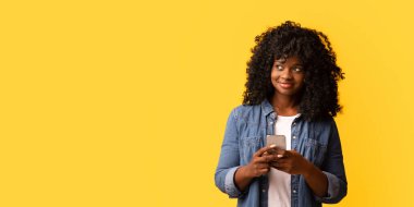 Gülümseyen kıvırcık Afrikalı Amerikalı genç kadın cep telefonuyla fotokopi odasına bakıyor, sarı arka plan. Güzel siyahi kadın elinde akıllı telefon tutuyor, yeni bir uygulama kullanıyor, reklamlara bakıyor.