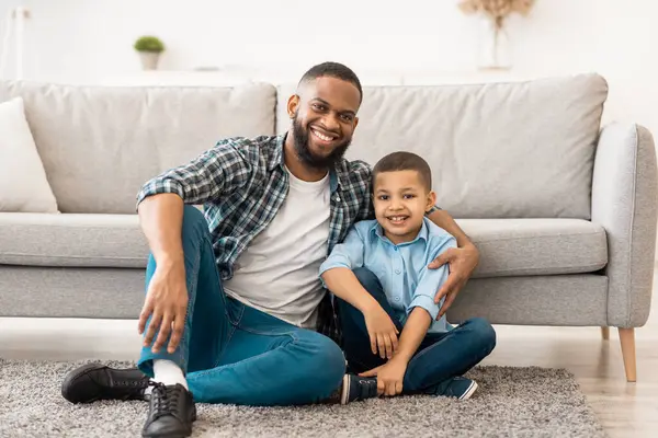 Mutlu Afrika kökenli Amerikalı baba ve oğul yerde oturmuş kameraya gülümsüyor. Genç baba ve çocuğunun portresi. Aile bağı, Mutluluk Konsepti