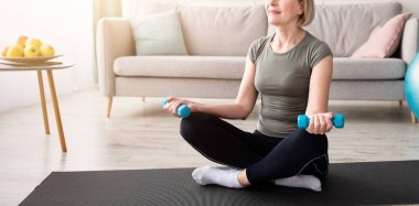 Spor giyim giymiş, halterli, yoga minderiyle egzersiz yapan sağlıklı, olgun bir kadın. Boş bir alan. Pozitif kıdemli bayan ağırlık çalışıyor, aile içi eğitim alıyor.