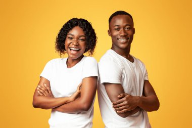 Başarılı Afrikalı Amerikalı erkek ve kadın kollarını göğsünde kavuşturarak sırt sırta verip turuncu stüdyonun arka planında kameraya gülümsüyor, neşe ve mutluluk gösteriyorlar.