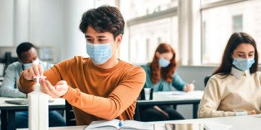 Temizlik ve Koruma Konsepti. Asyalı öğrenci şişeden elde antibakteriyel dezenfektan uyguluyor, ameliyat maskesi takıyor, sınıfta oturuyor. Enfeksiyon ve mikropların yayılmasını önler.